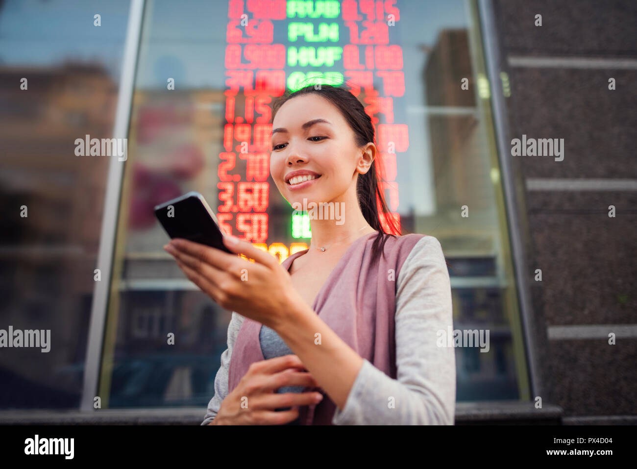 Una giovane bella donna asiatica utilizzando un'applicazione nel suo smart phone per controllare i tassi di cambio di fronte illuminata la scheda Informazioni. Foto Stock