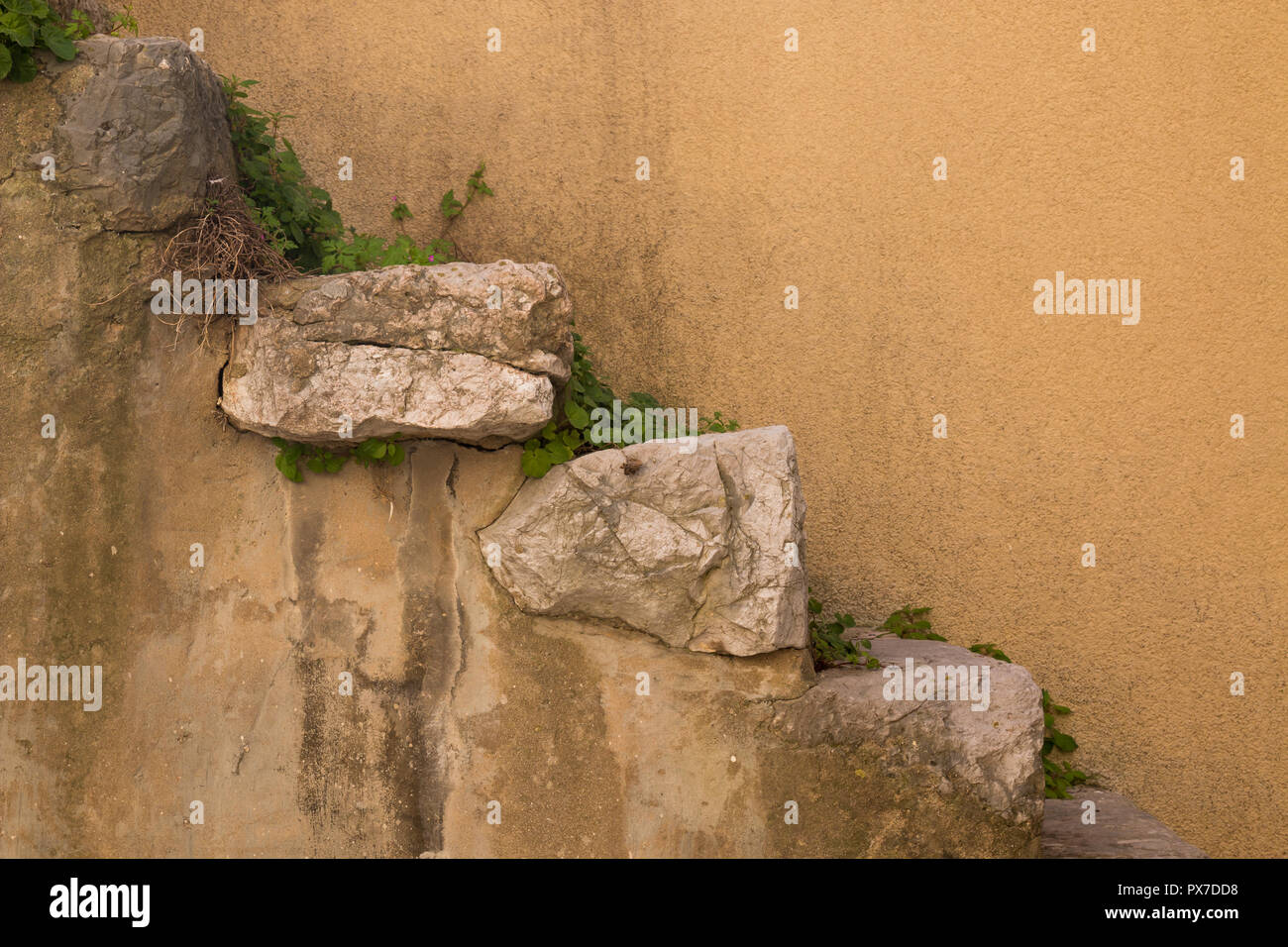 Muro di un edificio con una luce arancione facciata e scale fatte di grandi pezzi naturale di una pietra. Impianti tra le pietre. Città storica di Vrbnik, islan Foto Stock