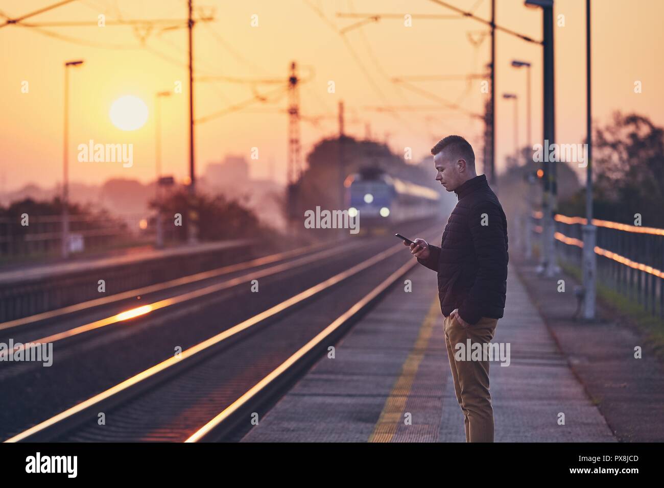 Giovane uomo in attesa presso la stazione ferroviaria di piattaforma e utilizzando smart phone contro il pendolarismo treno a sunrise. Foto Stock