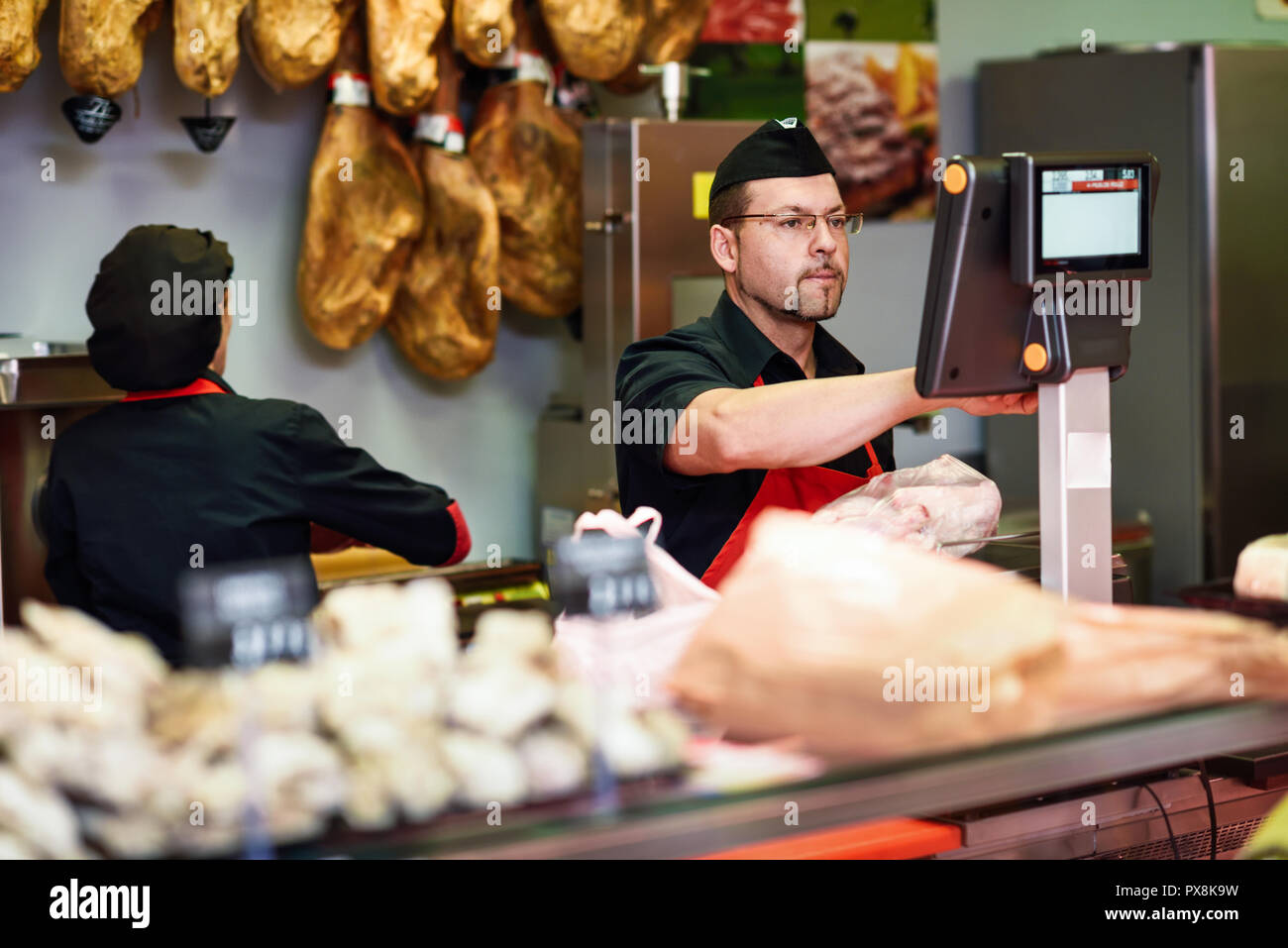 Macellaio in una macelleria di peso della carne e la carica con il prosciutto in background Foto Stock