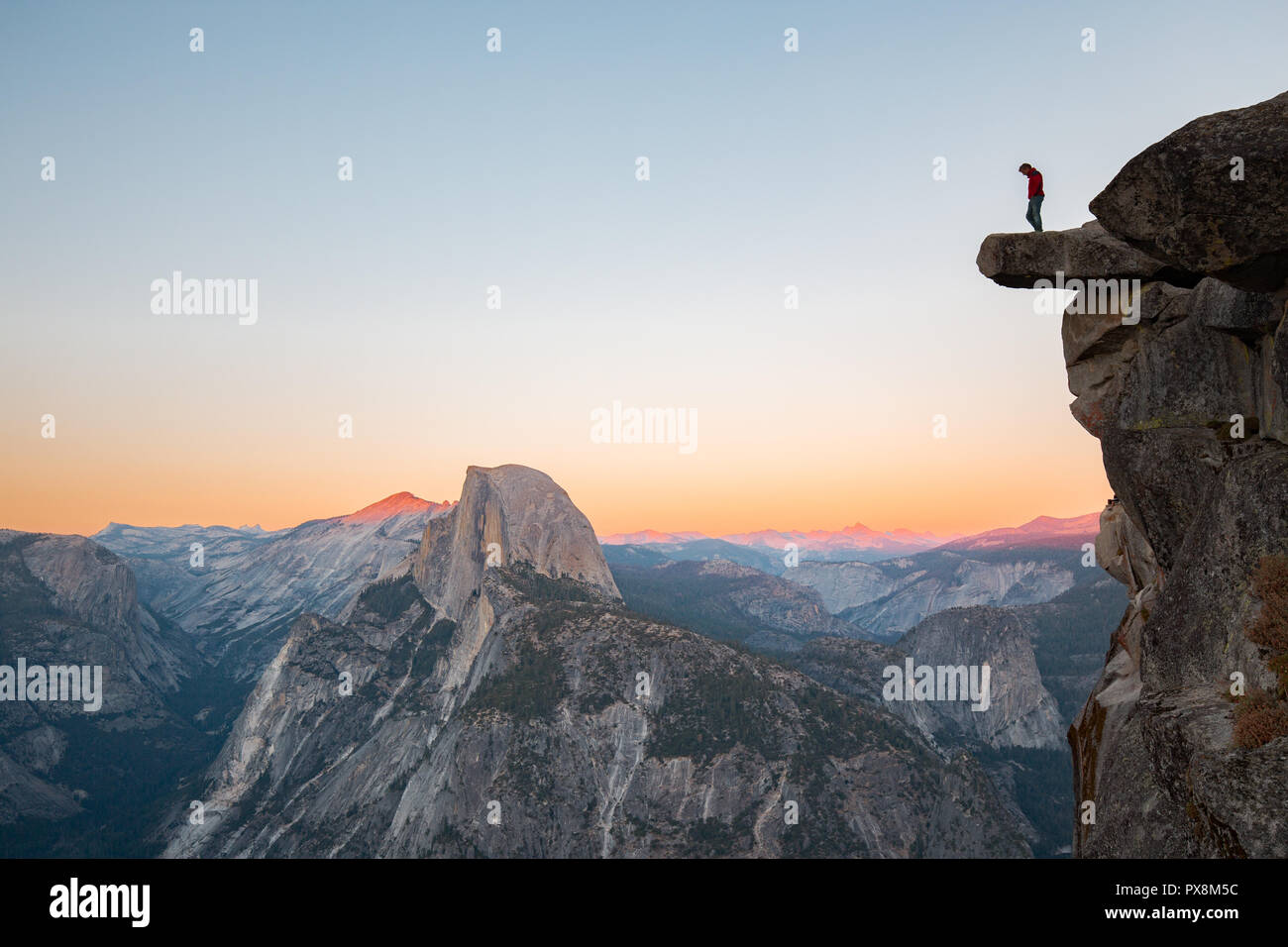 Un intrepido escursionista è in piedi su una roccia a strapiombo godendo della vista verso il famoso Half Dome presso il Glacier Point si affacciano nella bellissima crepuscolo serale Foto Stock