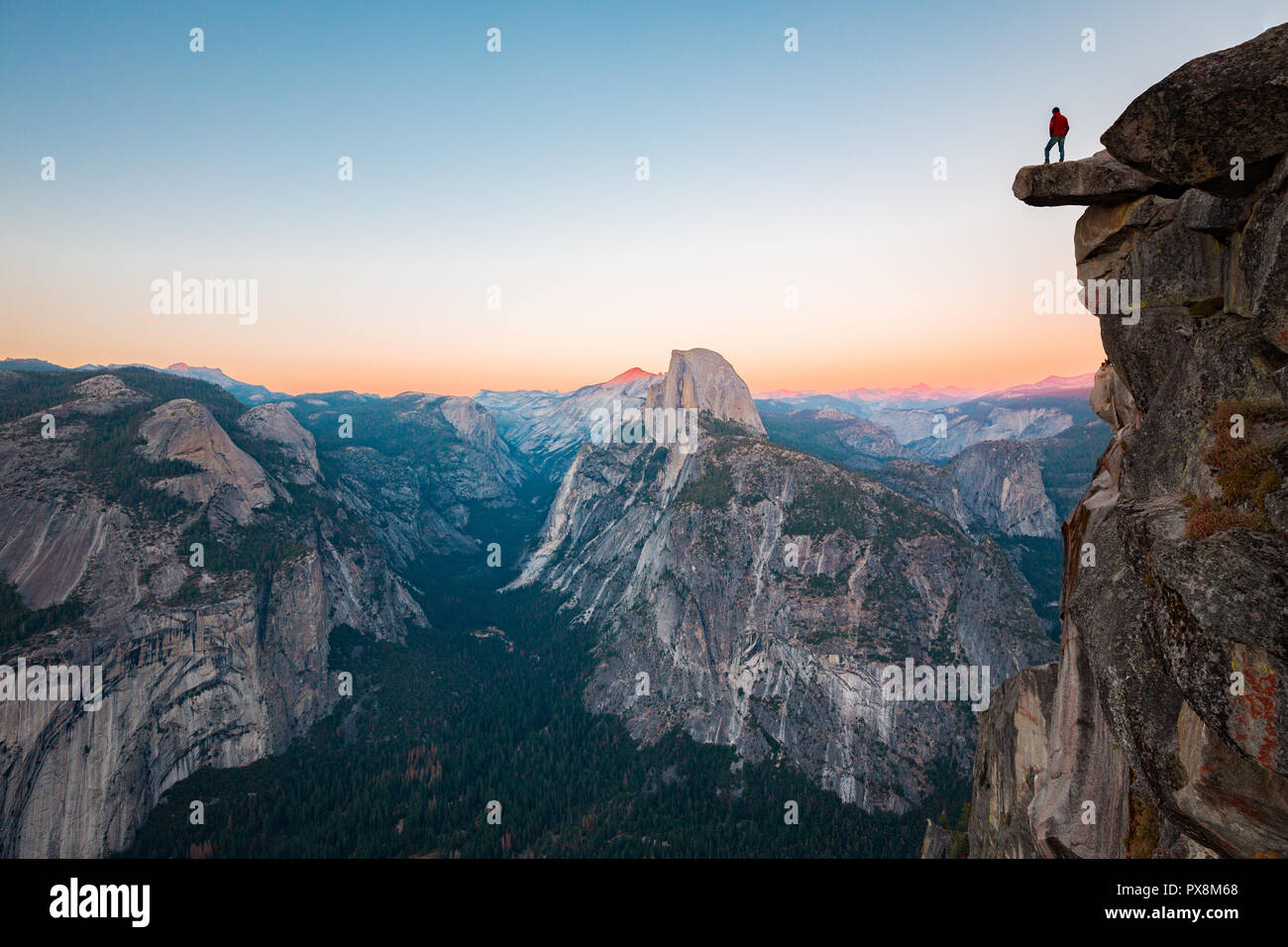 Un intrepido escursionista è in piedi su una roccia a strapiombo godendo della vista verso il famoso Half Dome presso il Glacier Point si affacciano nella bellissima crepuscolo serale Foto Stock