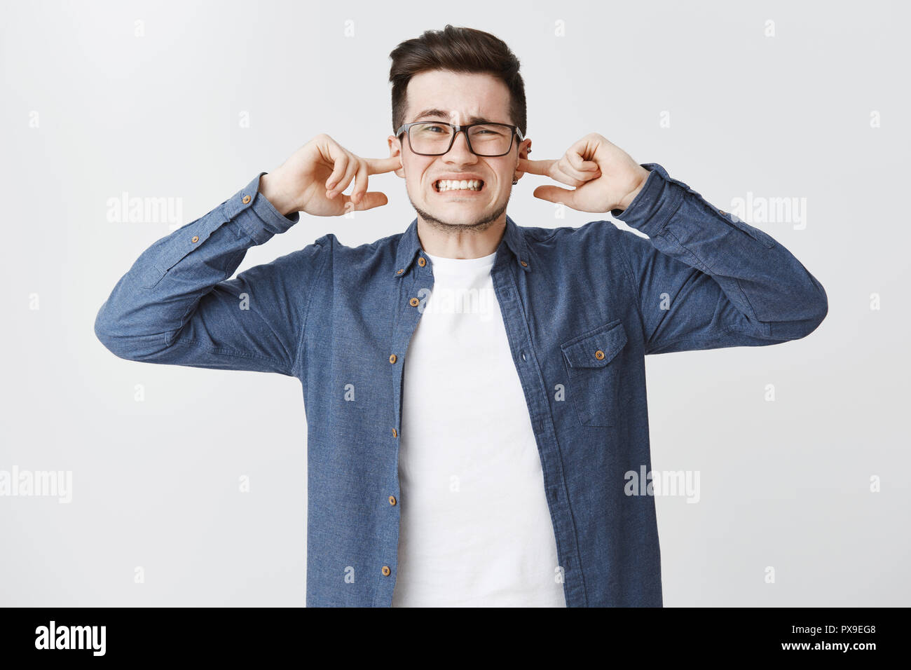 Infastidito e irritato sconvolto carino giovane uomo in bicchieri di denti di aggraffaggio da insoddisfazione accigliata chiudendo le orecchie con le dita indice essendo infastiditi con forte rumore terribile su sfondo bianco Foto Stock