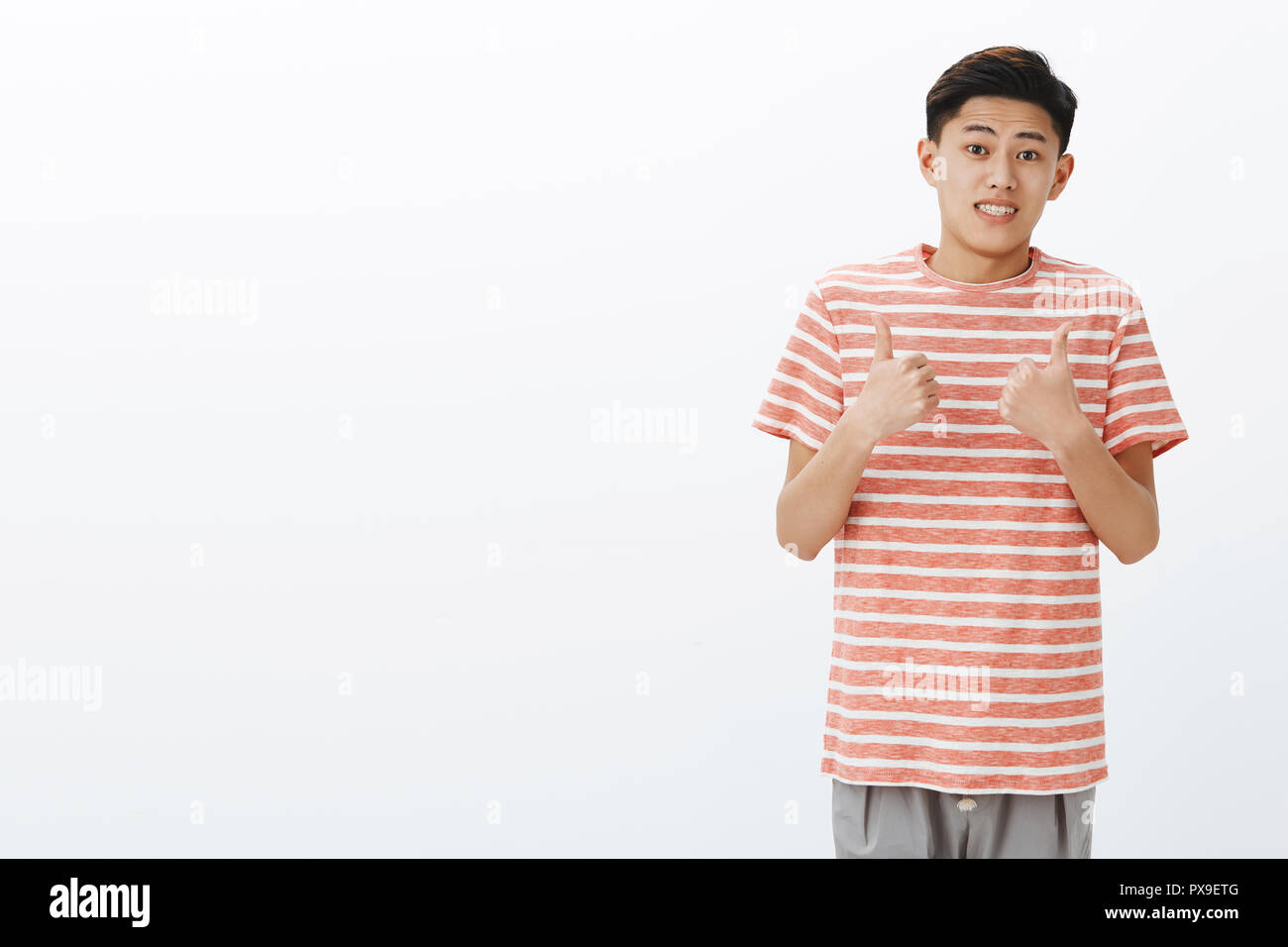Bene, immagino. Ritratto di sicuro scomoda attraente giovane uomo asiatico in striped t-shirt rendendo stretta ucertain sorriso e mostrando i pollici fino gesto come se accetta o come idea, in posa su uno sfondo grigio Foto Stock