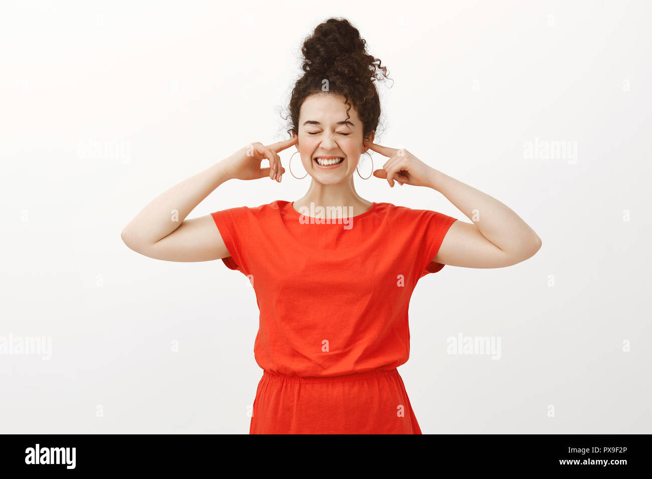 Ritratto di positivo bella donna in abito rosso, sorridente ampiamente e che copre le orecchie con le dita indice, in piedi con gli occhi chiusi, eccitato in attesa di bang da fuochi d' artificio su sfondo grigio Foto Stock