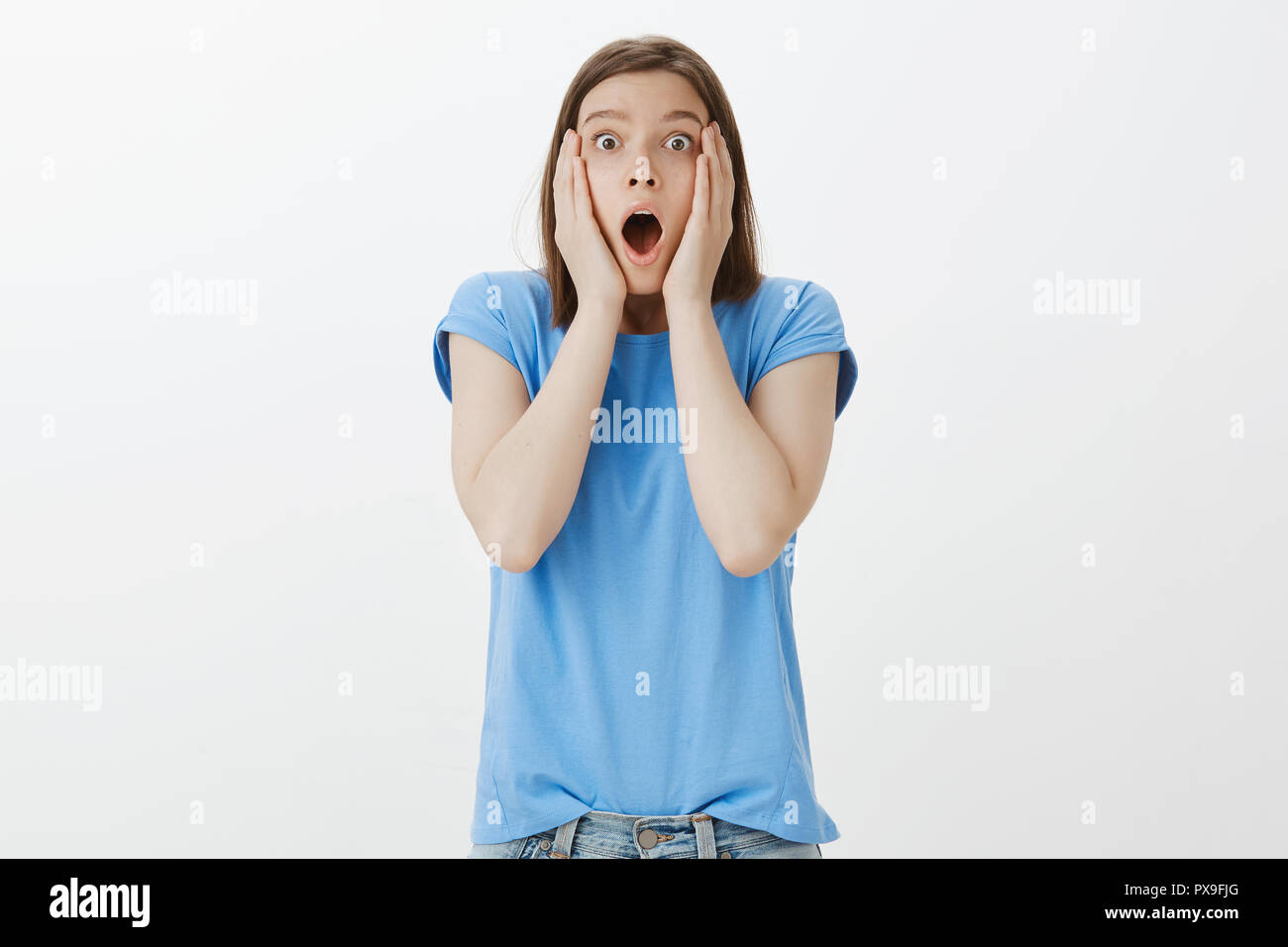 Alcun modo che non può accadere per un reale. Ritratto di impressionato e sconvolto divertente donna in blu t-shirt, la caduta della mascella, gridando e fissando con occhi schioccato in telecamera, tenendo le mani sul viso, storditi Foto Stock