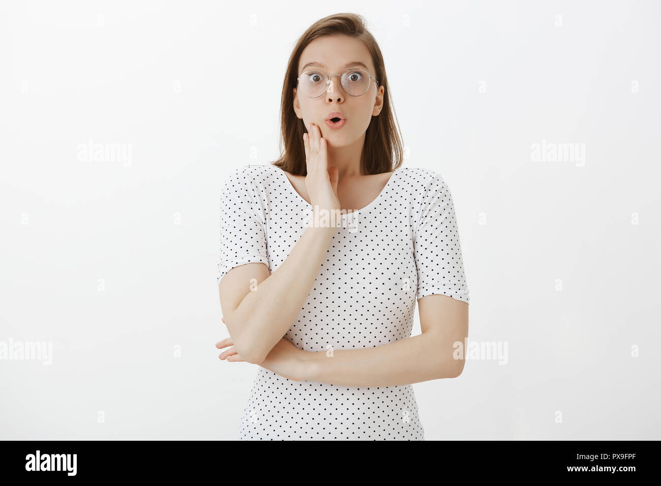 Inquadratura orizzontale di sconvolto e impressionato donna in bicchieri e alla moda di t-shirt, dicendo wow toccando il viso e a soffocare, fissando sorpreso in telecamera, udito incredibile incredibile rumor, divertito permanente Foto Stock