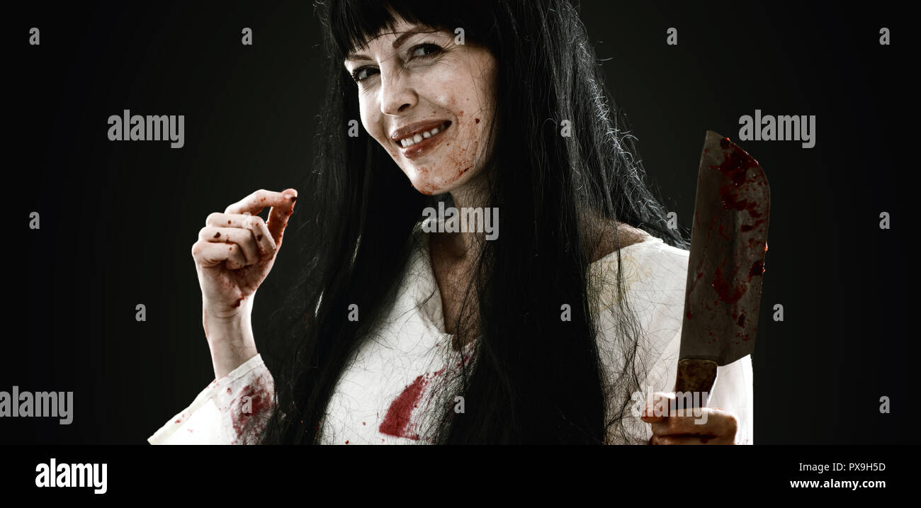 Halloween horror. Crazy sanguinosa spaventoso maniaco sorridente ragazza con il coltello macellaio cleaver guarda nella fotocamera su sfondo nero Foto Stock
