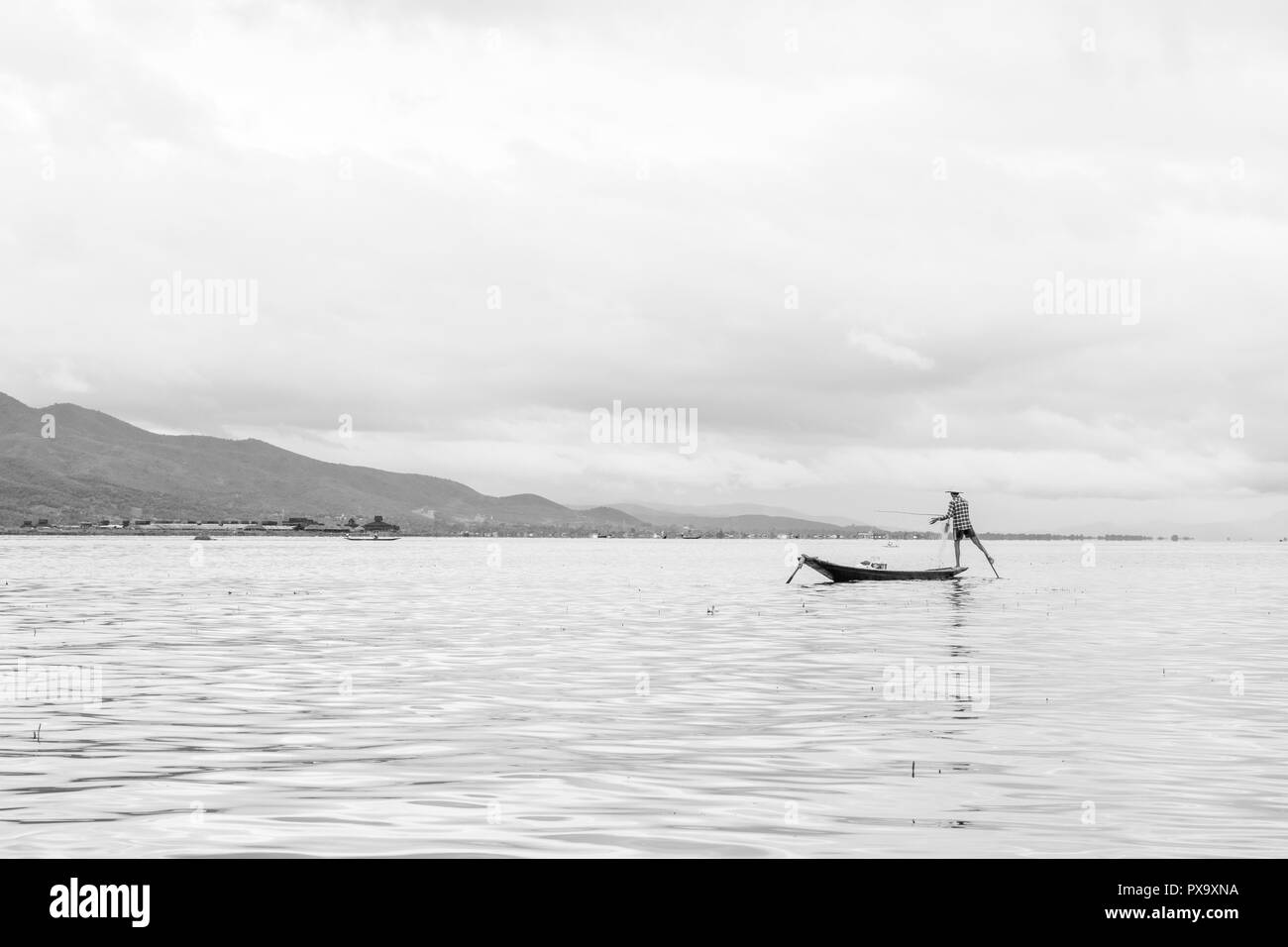 Locali di viaggio giovane maschio birmano pescatore controllato che indossa la maglietta, utilizzando la memory stick e net per pesci, in equilibrio su un piede sulla barca, Lago Inle MYANMAR Birmania Foto Stock