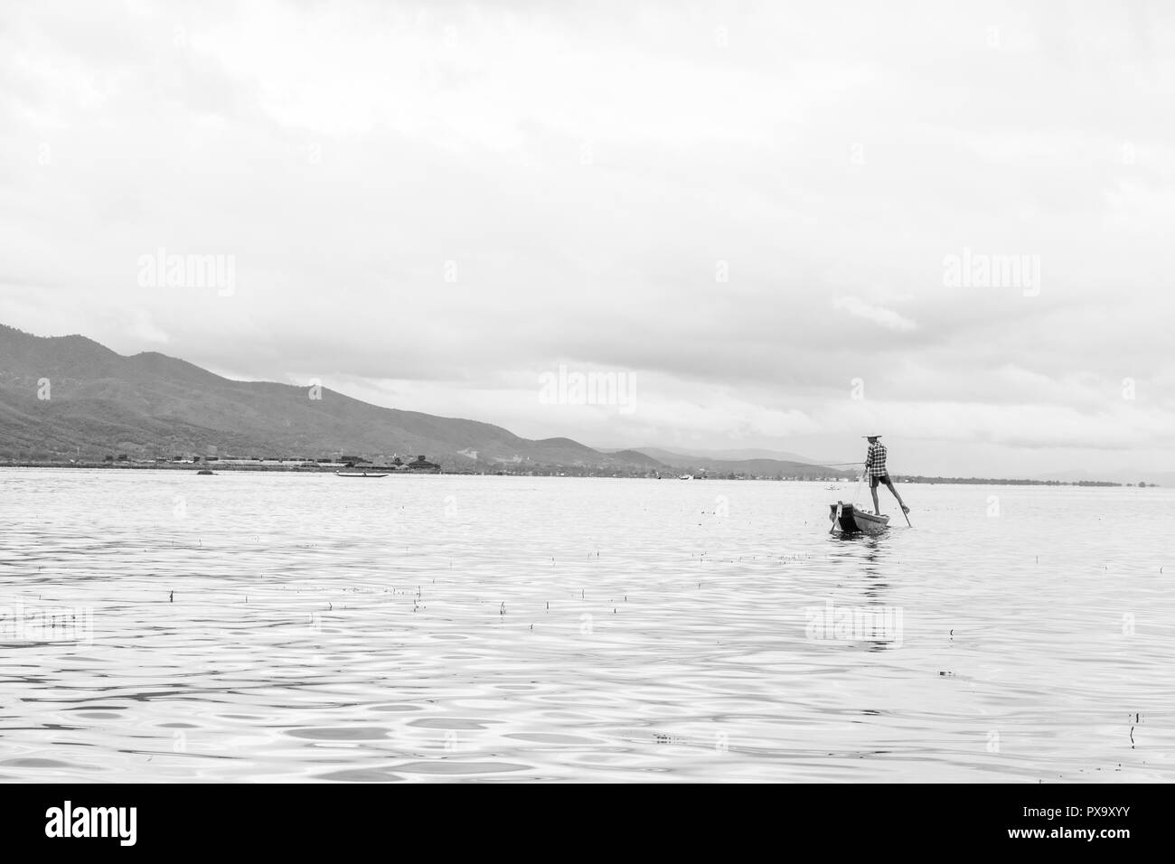Locali di viaggio giovane maschio birmano pescatore controllato che indossa la maglietta, utilizzando la memory stick e net per pesci, in equilibrio su un piede sulla barca, Lago Inle MYANMAR Birmania Foto Stock