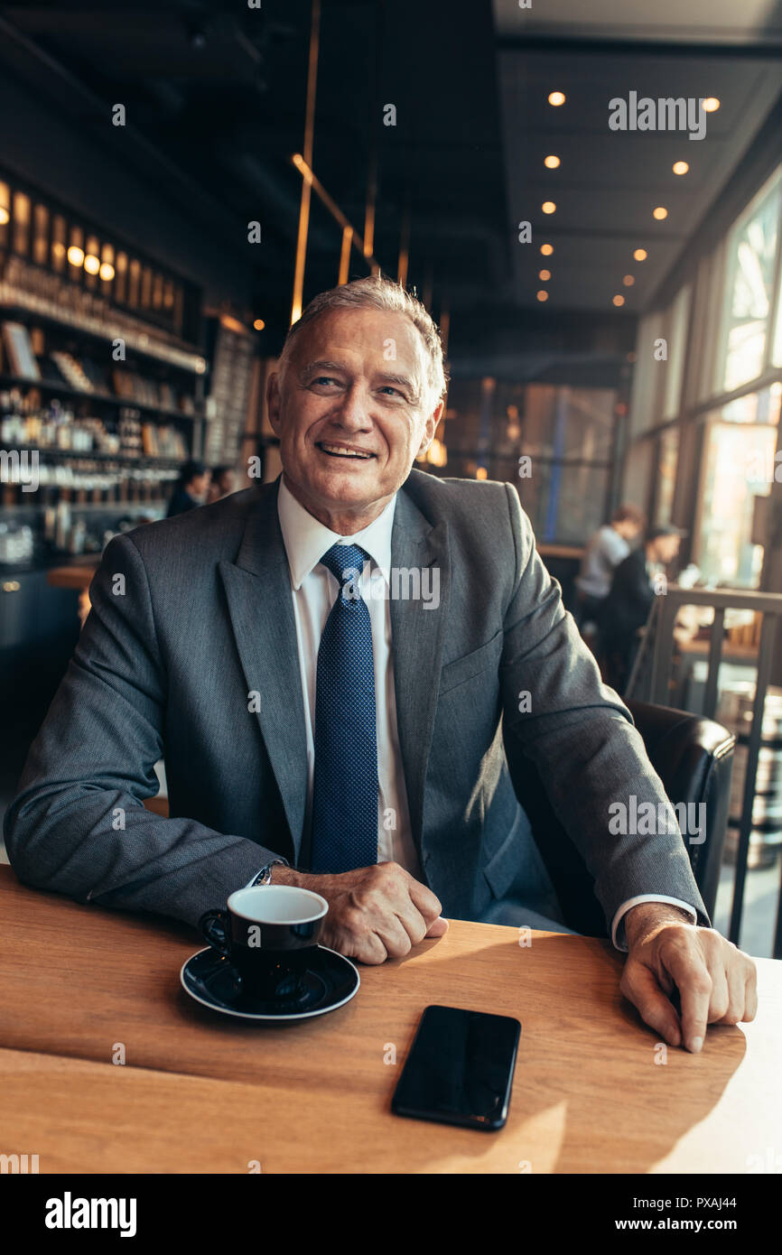 Ritratto di fiducia senior imprenditore seduto alla caffetteria moderna con tazza di caffè e telefono cellulare sul tavolo. Imprenditore maschio di relax presso il caffè sho Foto Stock