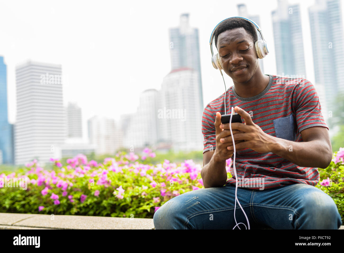 African uomo seduto in posizione di stazionamento mentre si ascolta la musica con le cuffie e utilizzando il telefono cellulare Foto Stock