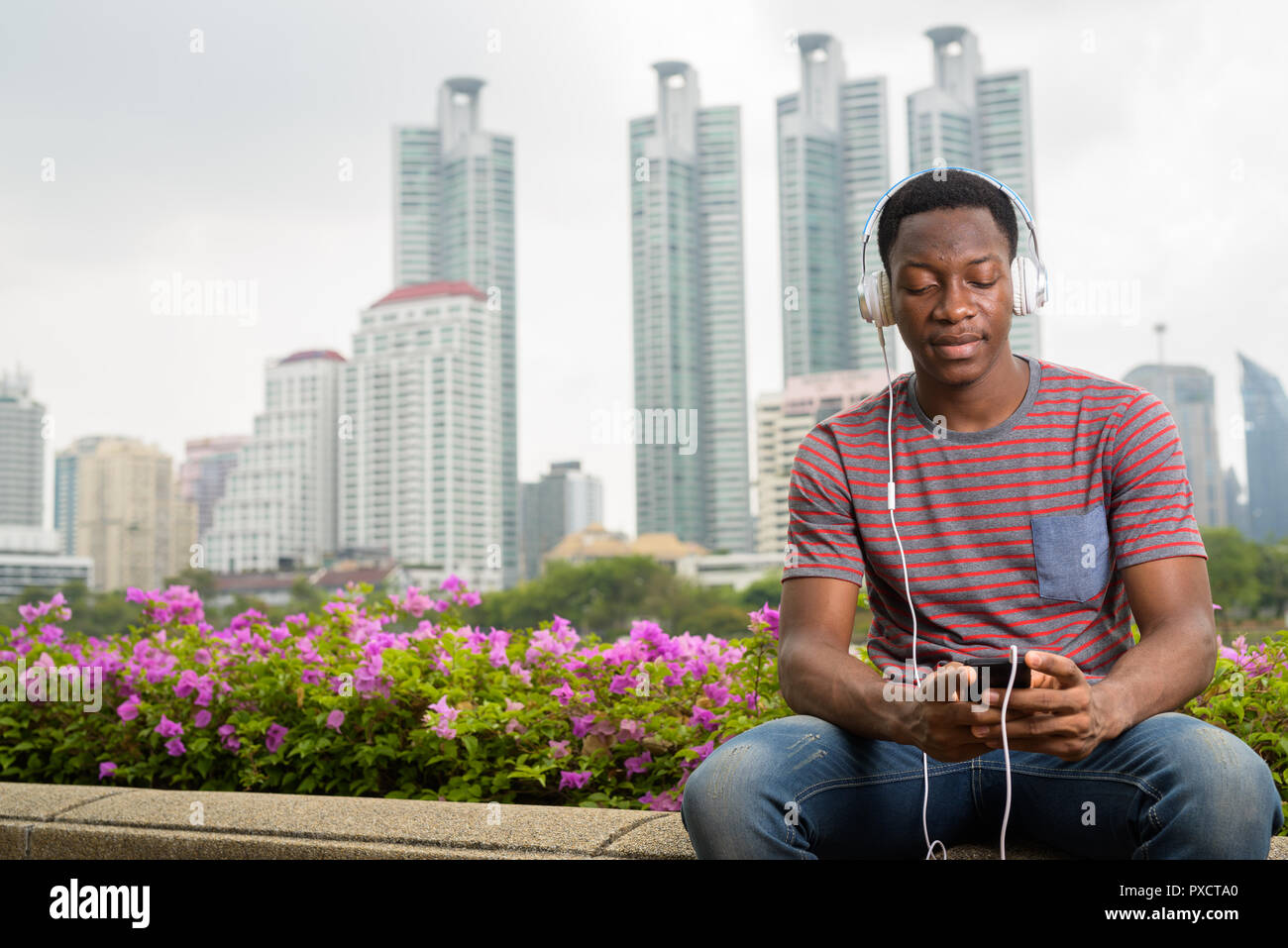 African uomo seduto in posizione di stazionamento mentre si ascolta la musica con le cuffie e utilizzando il telefono cellulare Foto Stock