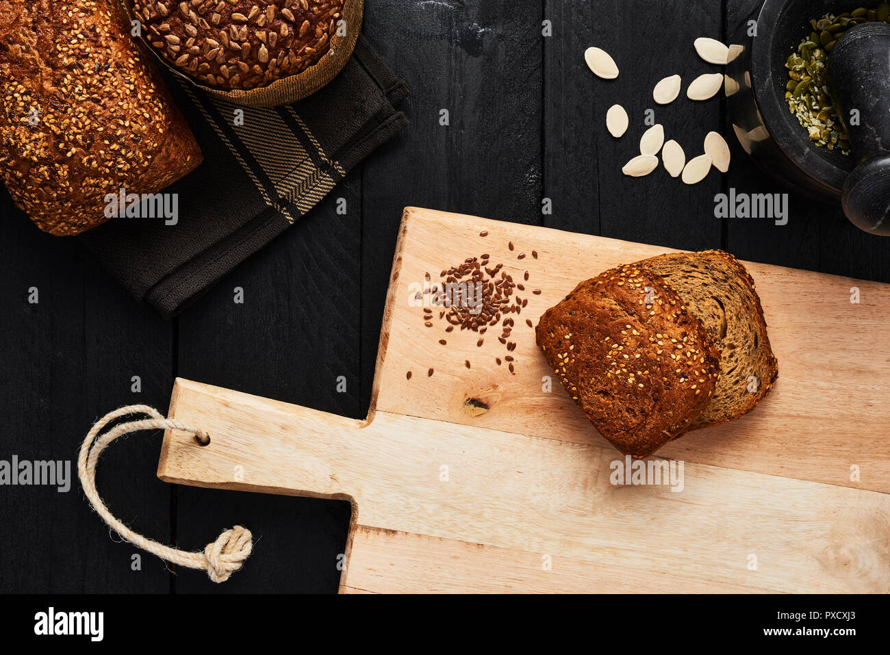 Tagliere con fette di pane, semi di lino e di vari crostini di pane integrale, semi di zucca e di mortaio su tavoli in legno nero. Vista dall'alto. Foto Stock