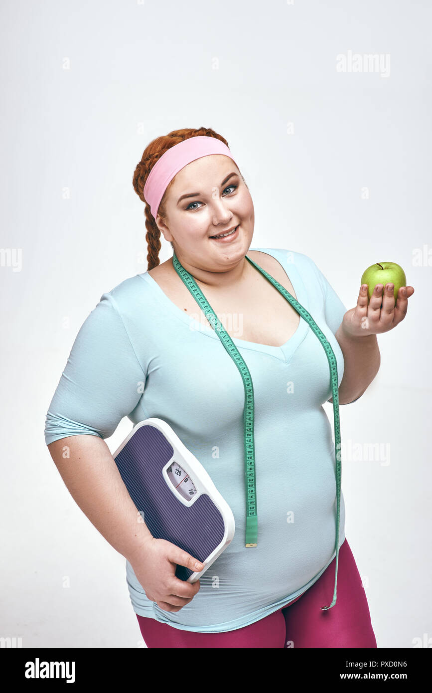 Divertente immagine di divertenti, dai capelli rossi, chubby donna su sfondo bianco. Donna che mantiene un apple e bilancia. Foto Stock