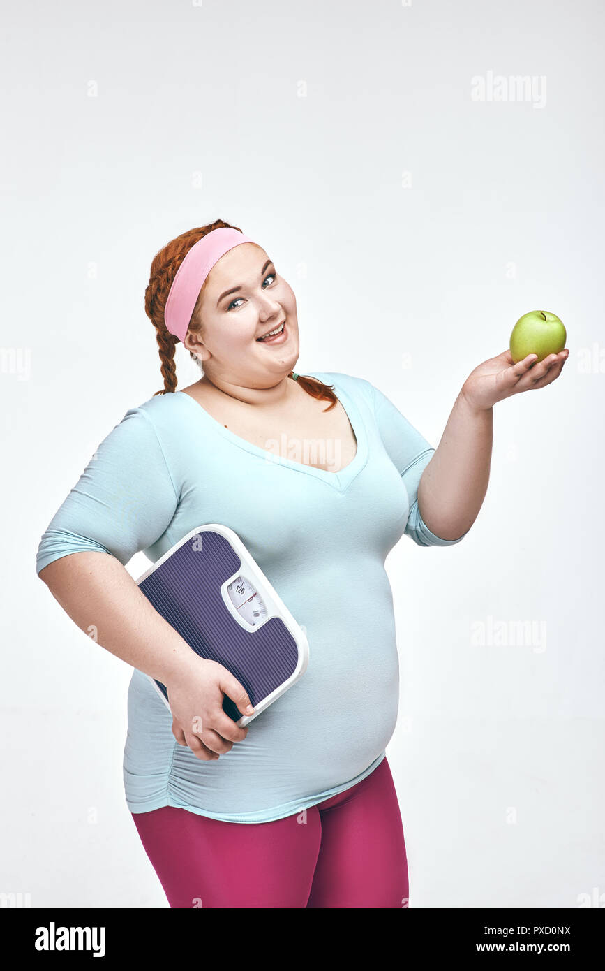 Divertente immagine di divertenti, dai capelli rossi, chubby donna su sfondo bianco. Donna che mantiene un apple e bilancia. Foto Stock
