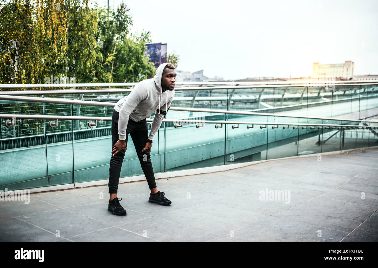 Giovane sportivo da uomo nero runner con gli auricolari e smartphone in una fascia da braccio sul ponte in una città di riposo. Copia dello spazio. Foto Stock