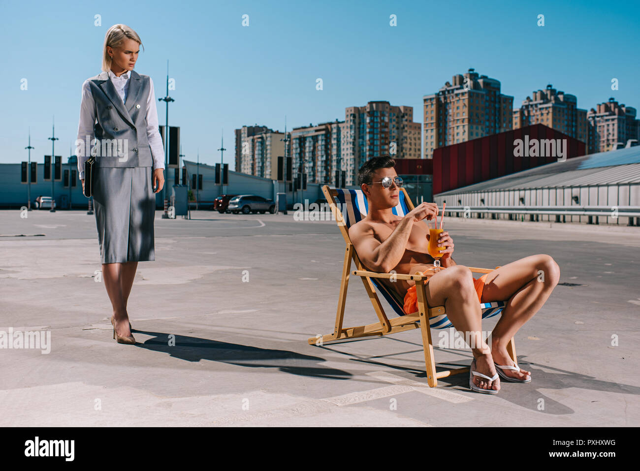 Bel giovane rilassante sulla sedia a sdraio mentre la sua signora boss in tuta in piedi accanto a lui su parcheggio Foto Stock
