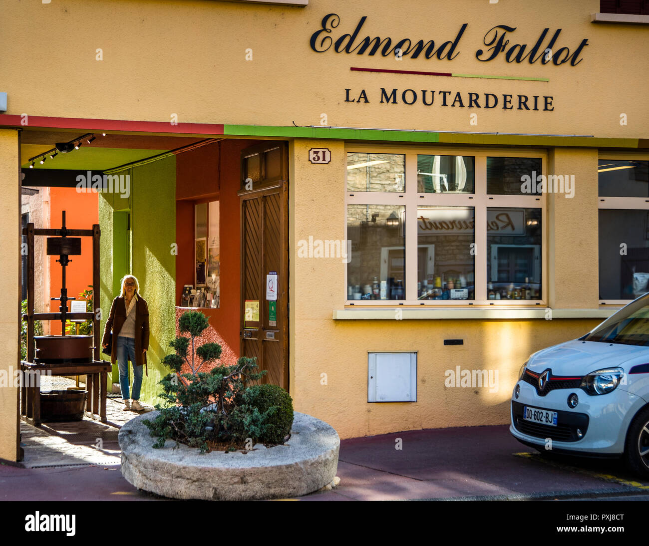 Un visitatore esce dall’arco della Moutarderie Edmond Fallot a Beaune in Borgogna, Francia, oltre una storica pressa olearia nell’area d’ingresso. Il produttore di senape produce in questo sito dal 1840 ed è oggi l’unico produttore di senape a gestione familiare rimasto in Francia. Foto Stock