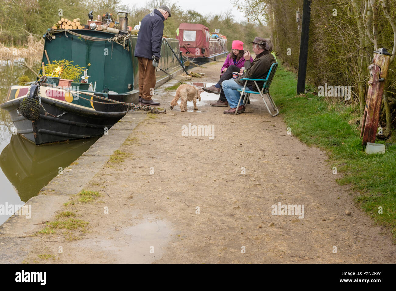 Persone sedute in sedie e bere il tè al fianco di un narrowboat ormeggiato sul canale. Un anziano uomo in piedi il suo cane ha un arresto di una sessione di conversazione testuale. Foxton. Foto Stock