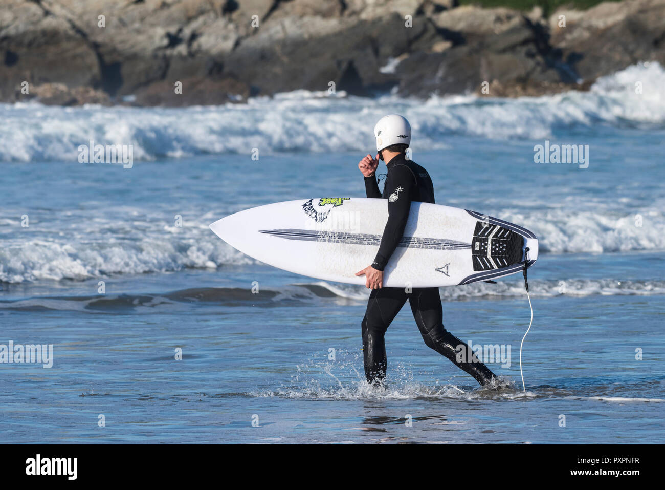 Protezione della testa - di un surfista che indossa un casco di sicurezza e portando la sua tavola da surf a piedi in mare a Fistral Beach in Newquay in Cornovaglia. Foto Stock