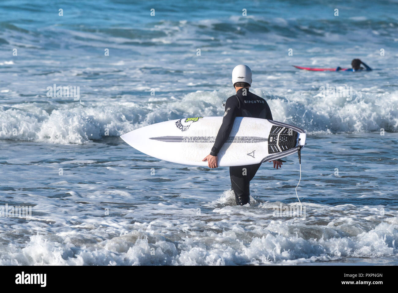 Un surfista che indossa un casco di sicurezza e portando la sua tavola da surf a piedi in mare a Fistral Beach in Newquay in Cornovaglia. Foto Stock