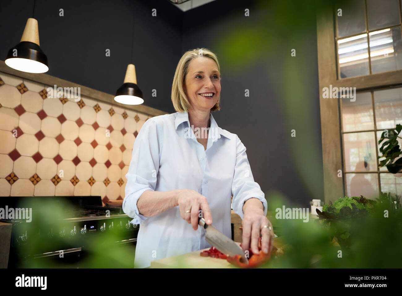 Sorridente donna matura tritare il peperone in cucina Foto Stock