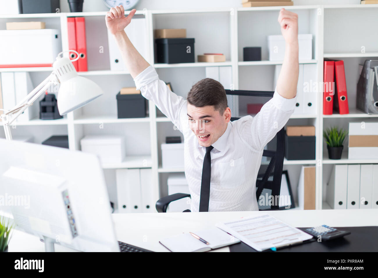Un giovane uomo seduto a una scrivania per computer in ufficio e ha sollevato le sue mani verso l'alto. Foto Stock
