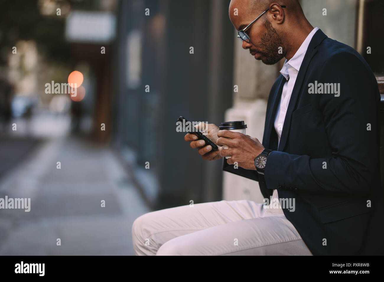 Vista laterale dell'uomo africano che indossa il costume seduto sulla strada scrivendo al telefono e tenendo in mano una tazza di caffè. uomo d'affari che si rilassa all'aperto con il telefono e. Foto Stock