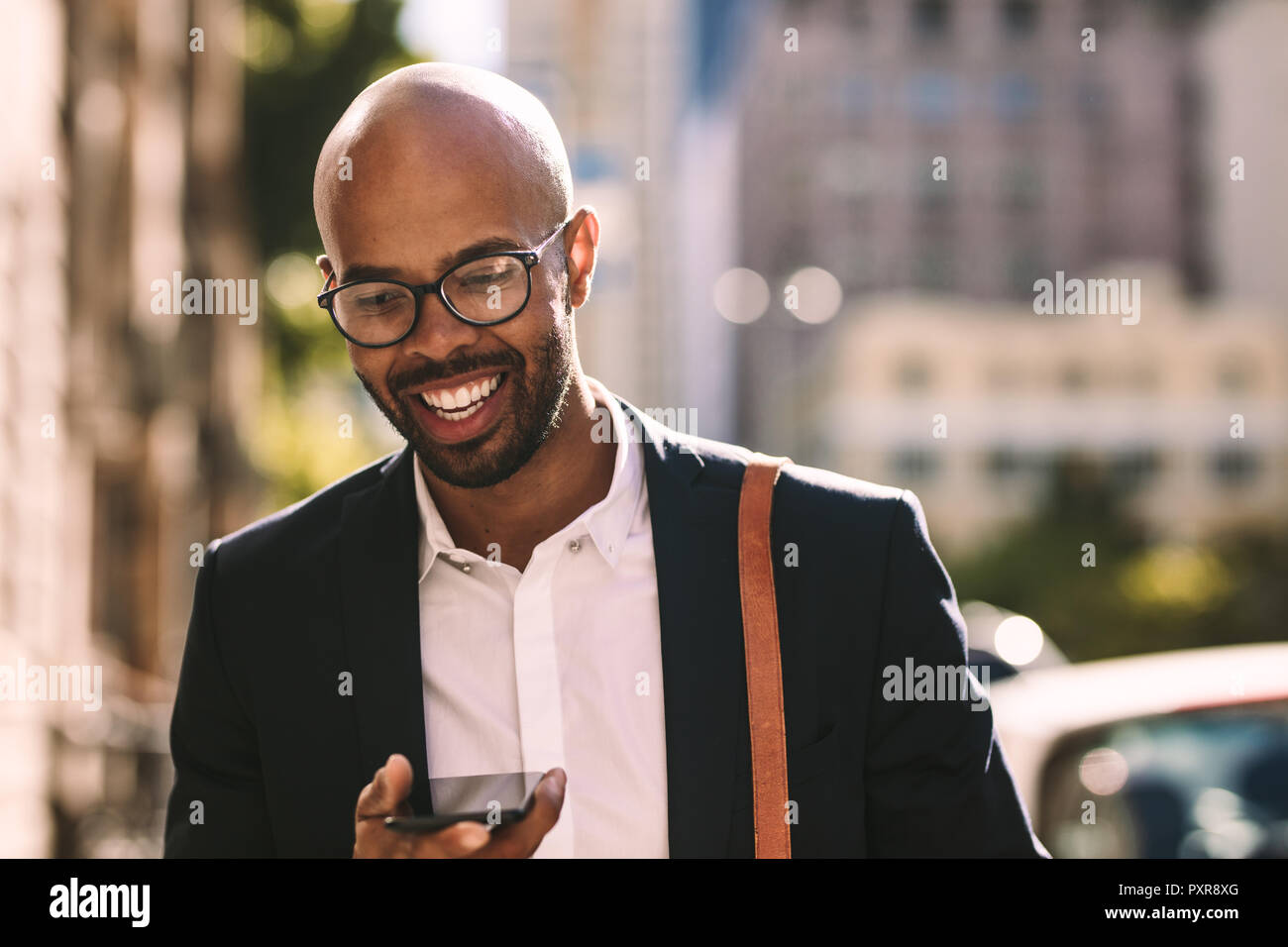 Sorridente giovane imprenditore africano il pendolarismo con un telefono cellulare mentre passeggiate all'aperto. Uomo calvo in tuta a piedi in città e parlando al cellulare Foto Stock