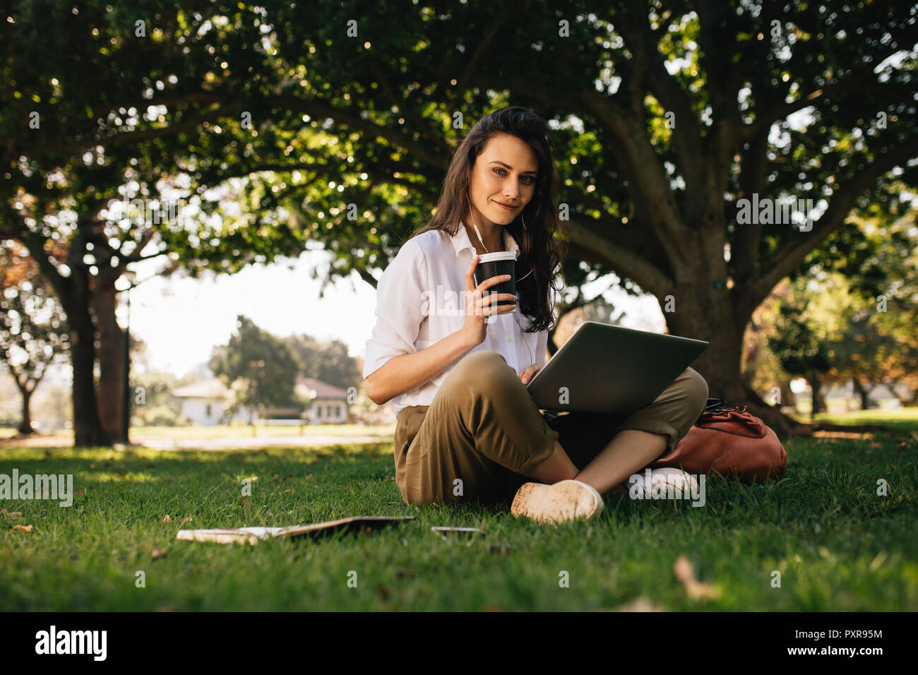 Bella donna seduta al parco con un caffè e un computer portatile. Femmina rilassante nel parco. Foto Stock