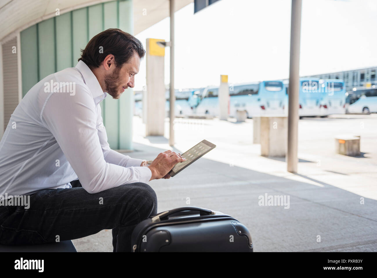 Imprenditore sorridente con la valigia seduto al bus terminal utilizzando tablet Foto Stock