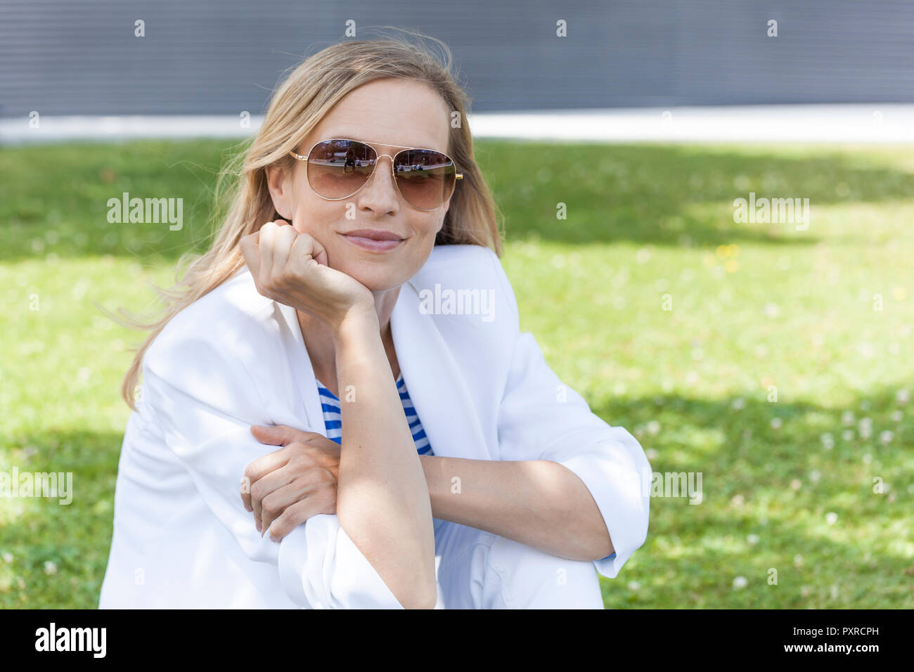 Sorridente bionda imprenditrice seduta sul prato, mano sul mento Foto Stock