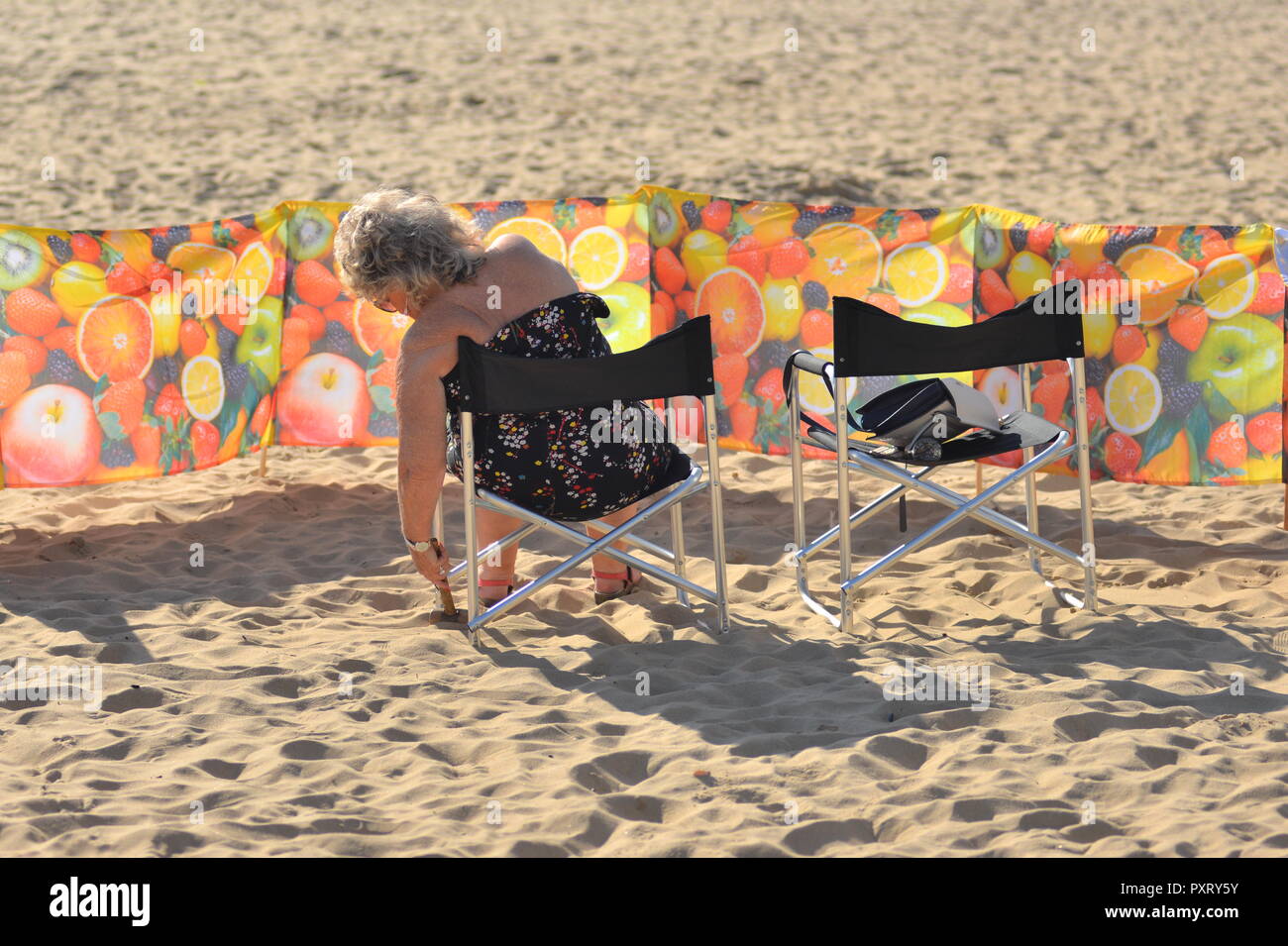 Donna seduta a prendere il sole in una sedia da spiaggia protetta da un parabrezza a motivi floreali, Boscombe, Bournemouth, Dorset, UK, 24th ottobre 2018, tempo: Caldo sole d'autunno. Foto Stock