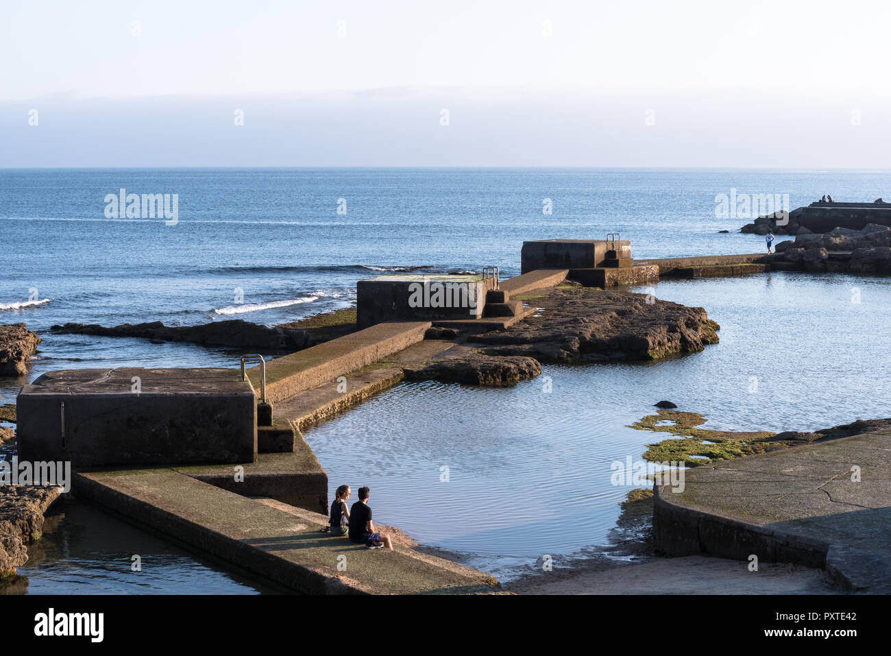 Estoril, Portogallo - 9 Luglio 2018: Coppia giovane seduto sui passi concreti della piscina di acqua di mare accanto al mare da parte di Boardwalk a Estoril Portogallo Foto Stock
