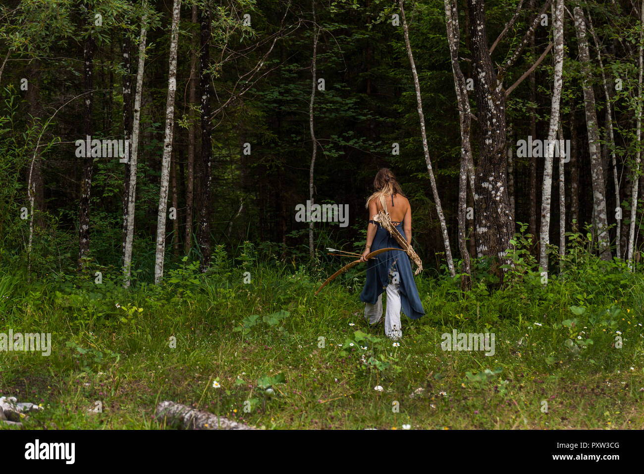 Vista posteriore della donna che cammina in una foresta con arco e frecce Foto Stock