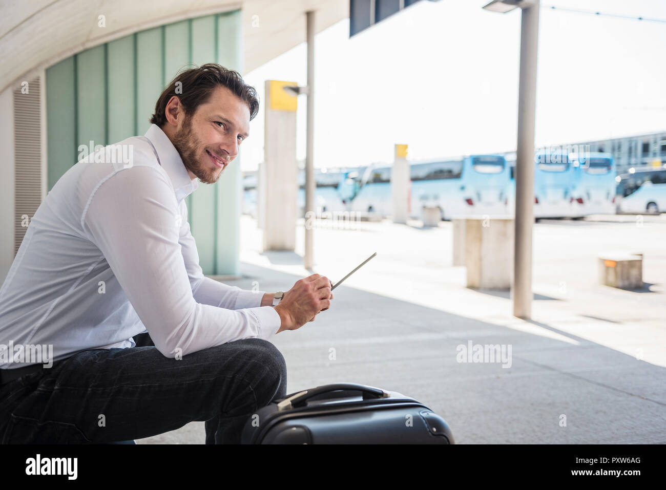 Imprenditore sorridente con la valigia seduto al bus terminal utilizzando tablet Foto Stock