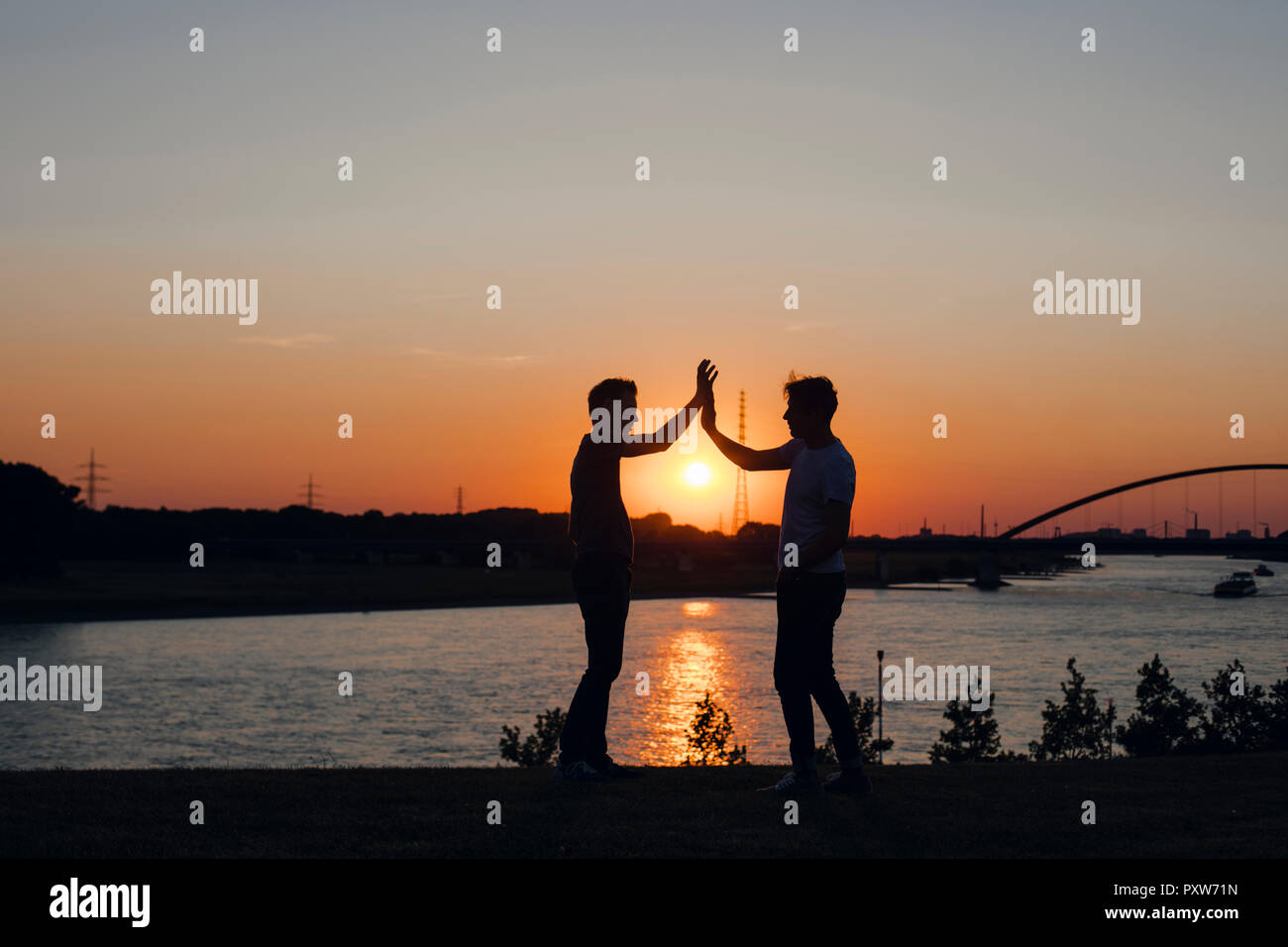 Due amici di alta fiving al tramonto, in piedi dal fiume Foto Stock