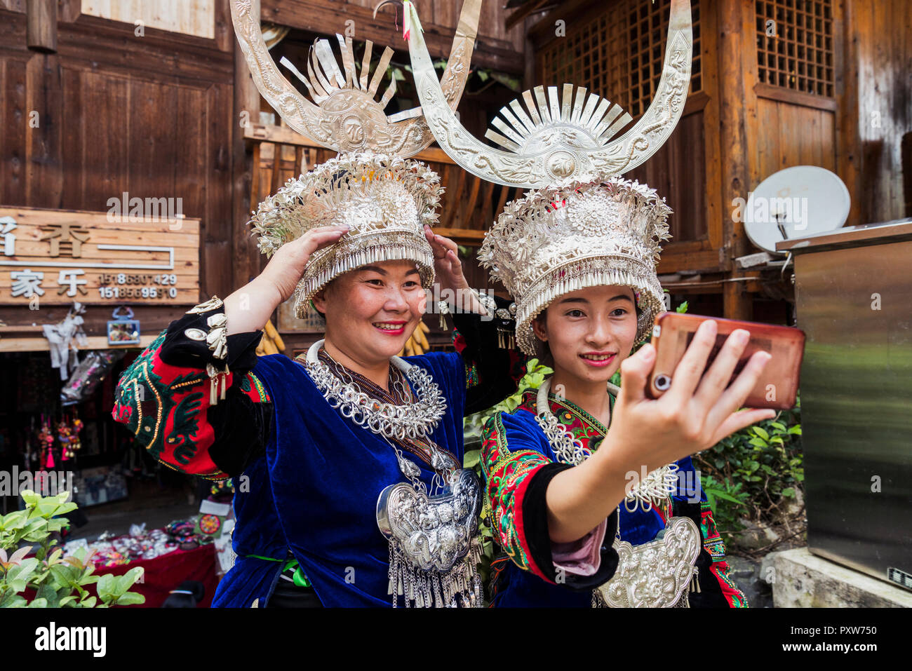 Cina, Guizhou, due Miao donne che indossano abiti tradizionali e copricapo prendendo un selfie con lo smartphone Foto Stock