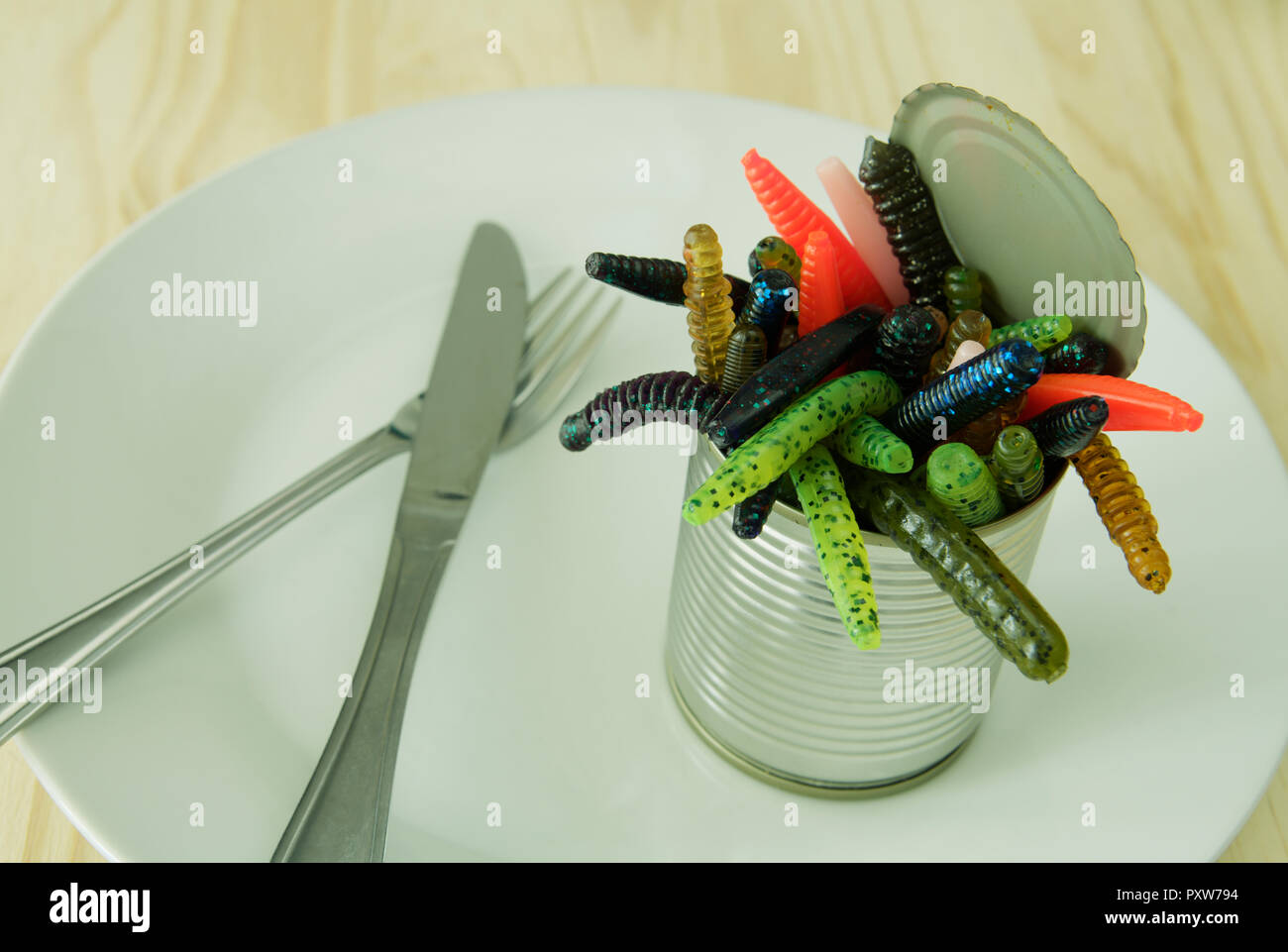 Situazione di vita di colorati strani vermi artificiale strisciando fuori del barattolo di latta sulla piastra di alimentare con coltello e forchetta Foto Stock