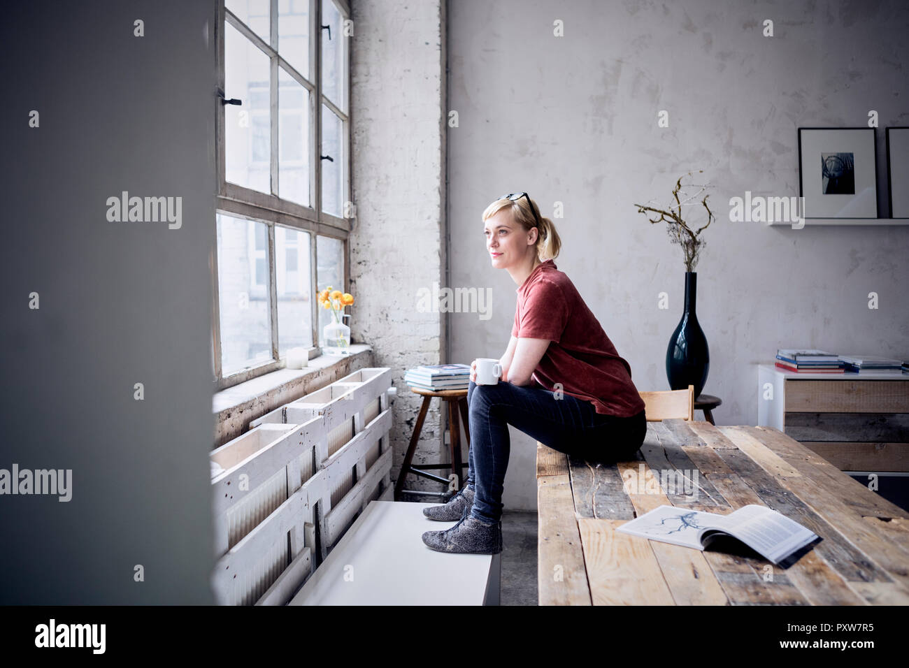 Donna sorridente con tazza da caffè seduti sulla scrivania in loft guardando attraverso la finestra Foto Stock
