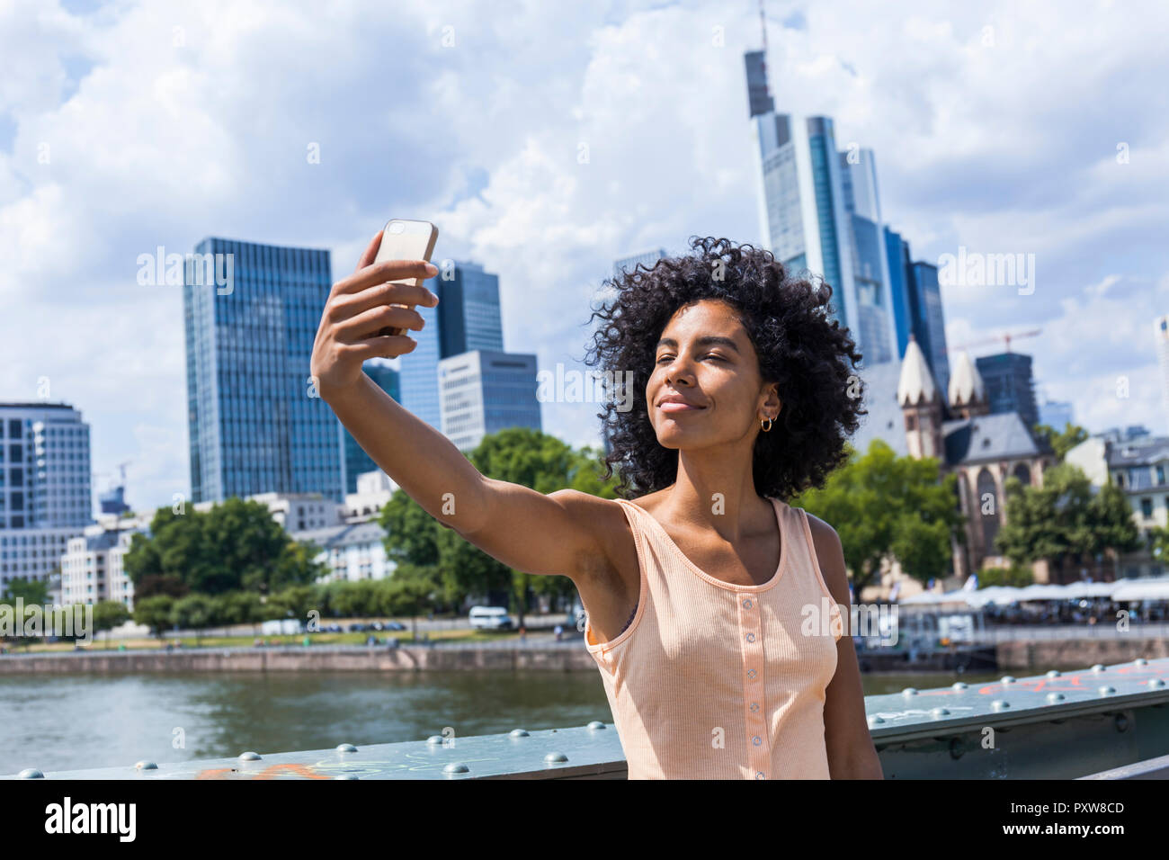 Germania, Francoforte, ritratto di contenuto giovane donna con capelli ricci tenendo selfie nella parte anteriore della skyline Foto Stock