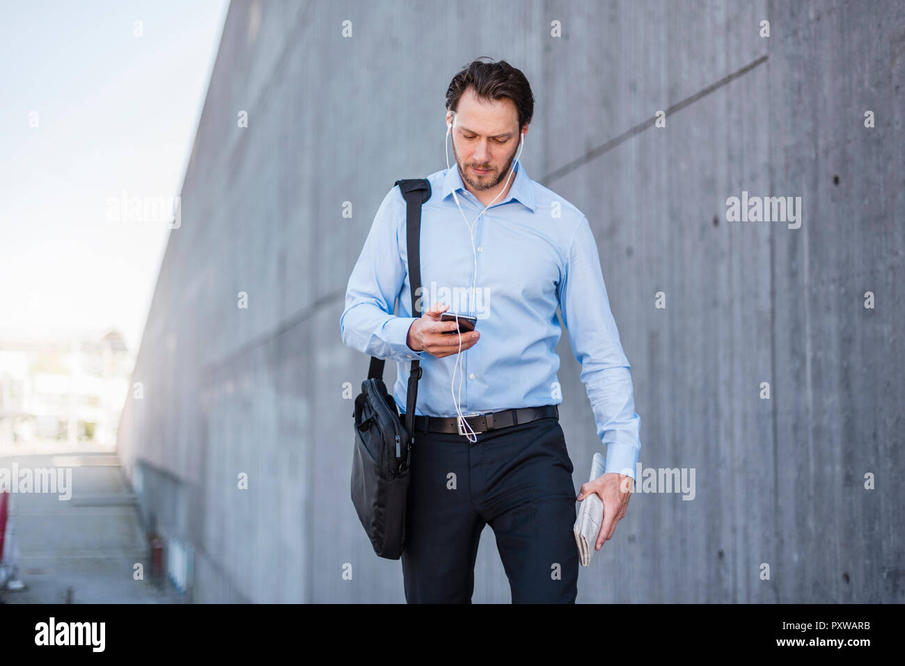 Imprenditore con gli auricolari camminando lungo il muro di cemento guardando smartphone Foto Stock