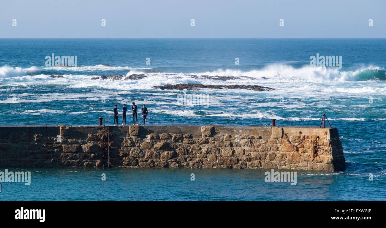 Vista in lontananza quattro uomini in abiti bagnati in piedi sul coperchio Sennen harbour parete affacciata sul mare a la massiccia le onde che si infrangono sulle rocce Foto Stock