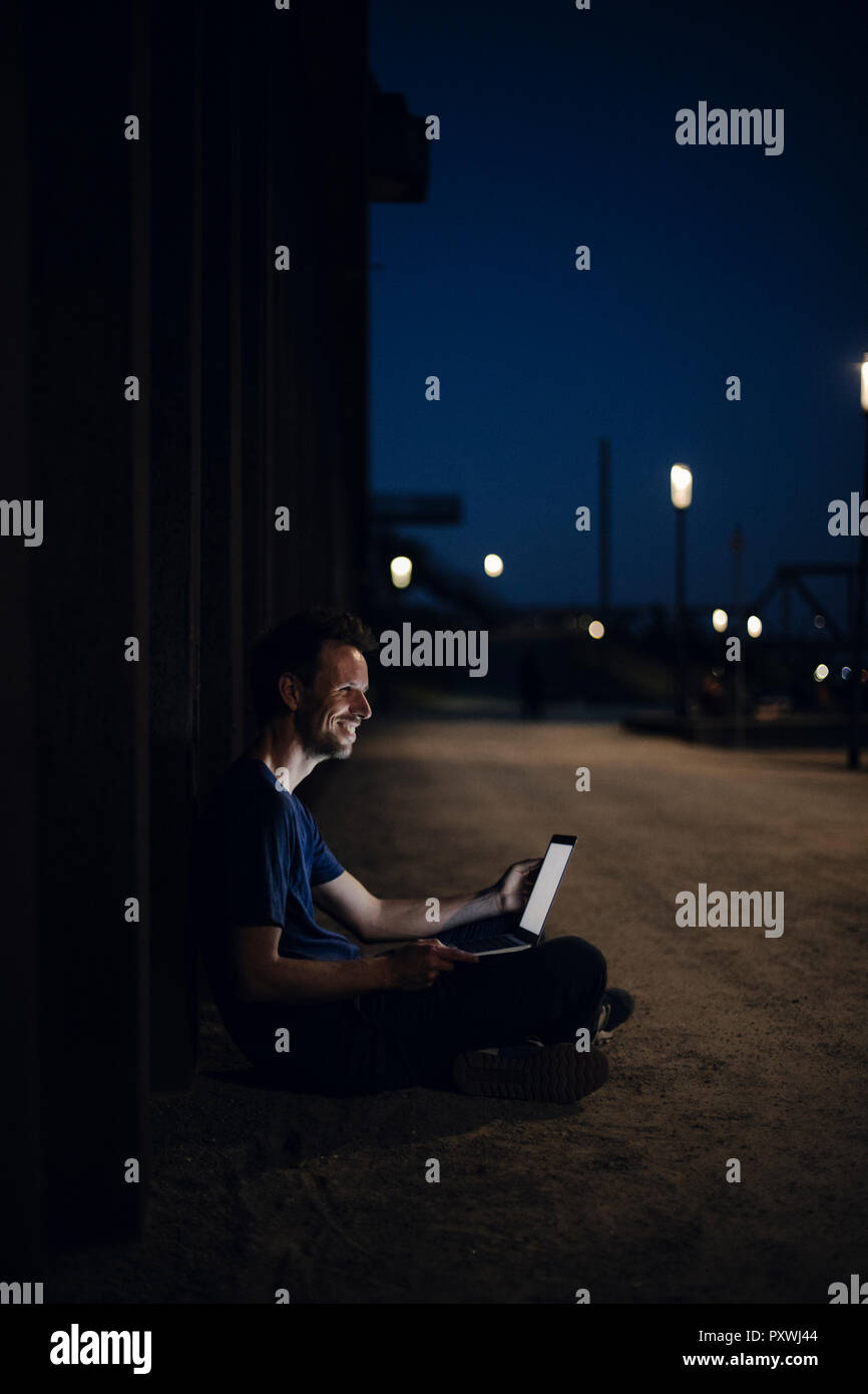 Metà uomo adulto seduto a gambe incrociate sulla terra, utilizzando computer portatile durante la notte Foto Stock