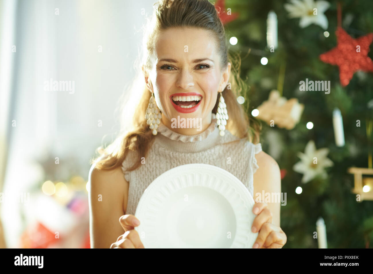 Sorridente giovane casalinga vicino albero di Natale azienda che serve cena bianco piastra Foto Stock