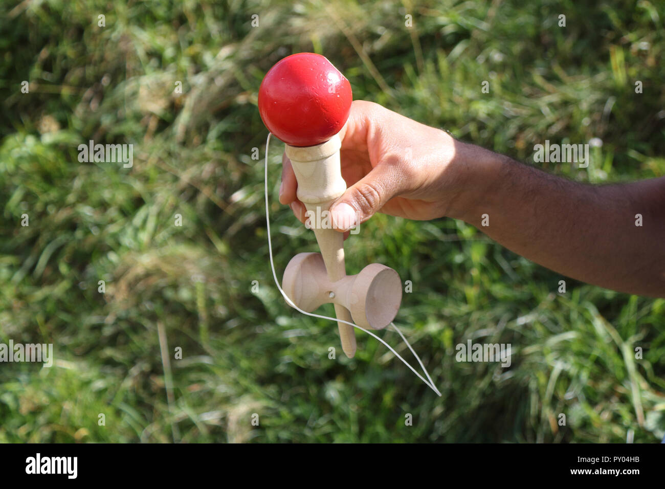 Un giovane ragazzo con un colore blu t-shirt tenendo un legno stick kendama e giocando con una palla rossa in un verde prato durante una soleggiata estate Foto Stock