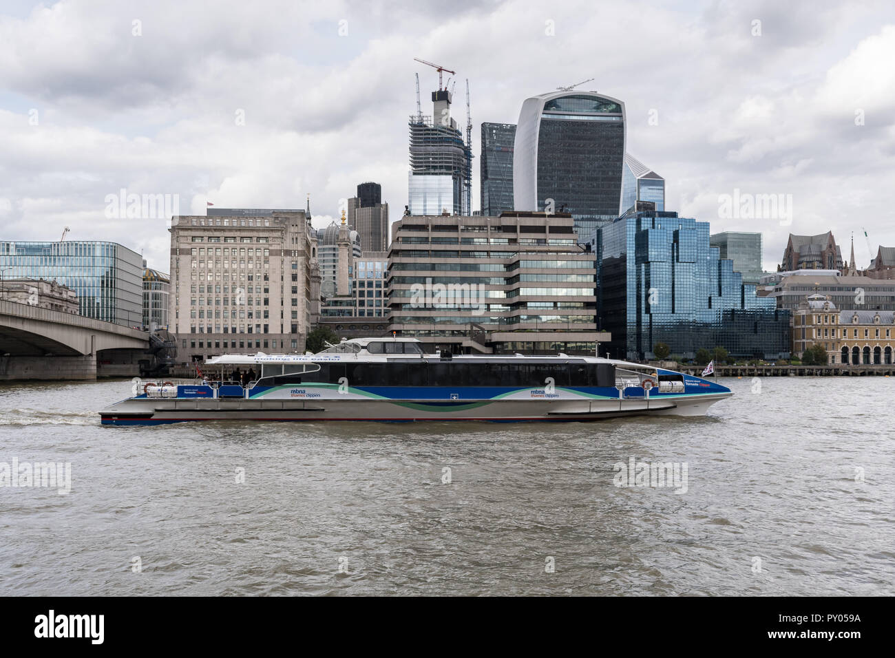 Una classe Tornado Thames clipper vele passato la città di Londra edifici lungo il Tamigi, London, Regno Unito Foto Stock
