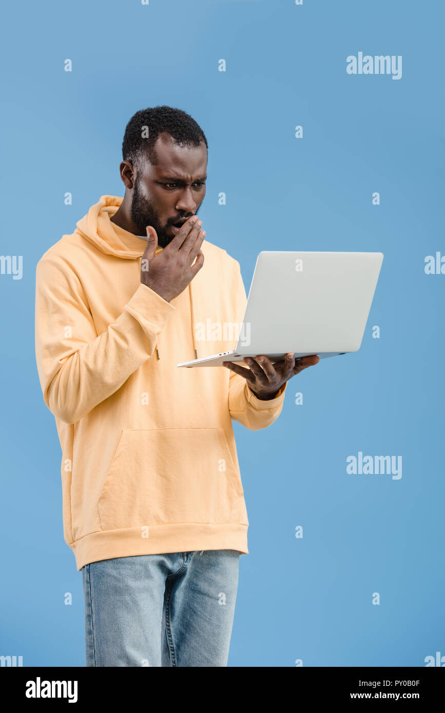 Scioccato African American uomo chiudendo la bocca con la mano e guardando il computer portatile isolato su sfondo blu Foto Stock
