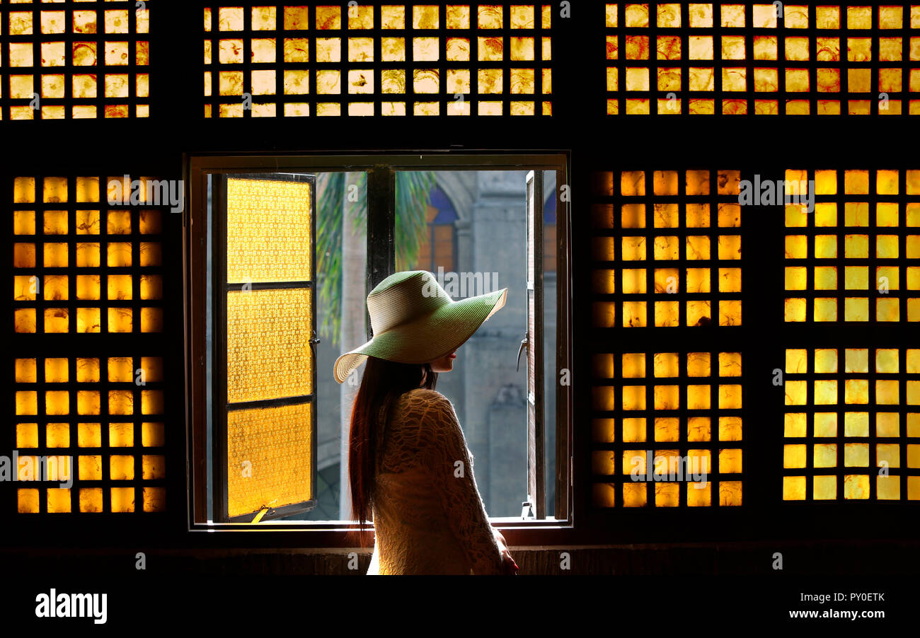 La donna in bianco Abito in pizzo e cappello nel corridoio di fronte capiz (shell) finestra in San Agustin Chiesa, Manila, Filippine Foto Stock